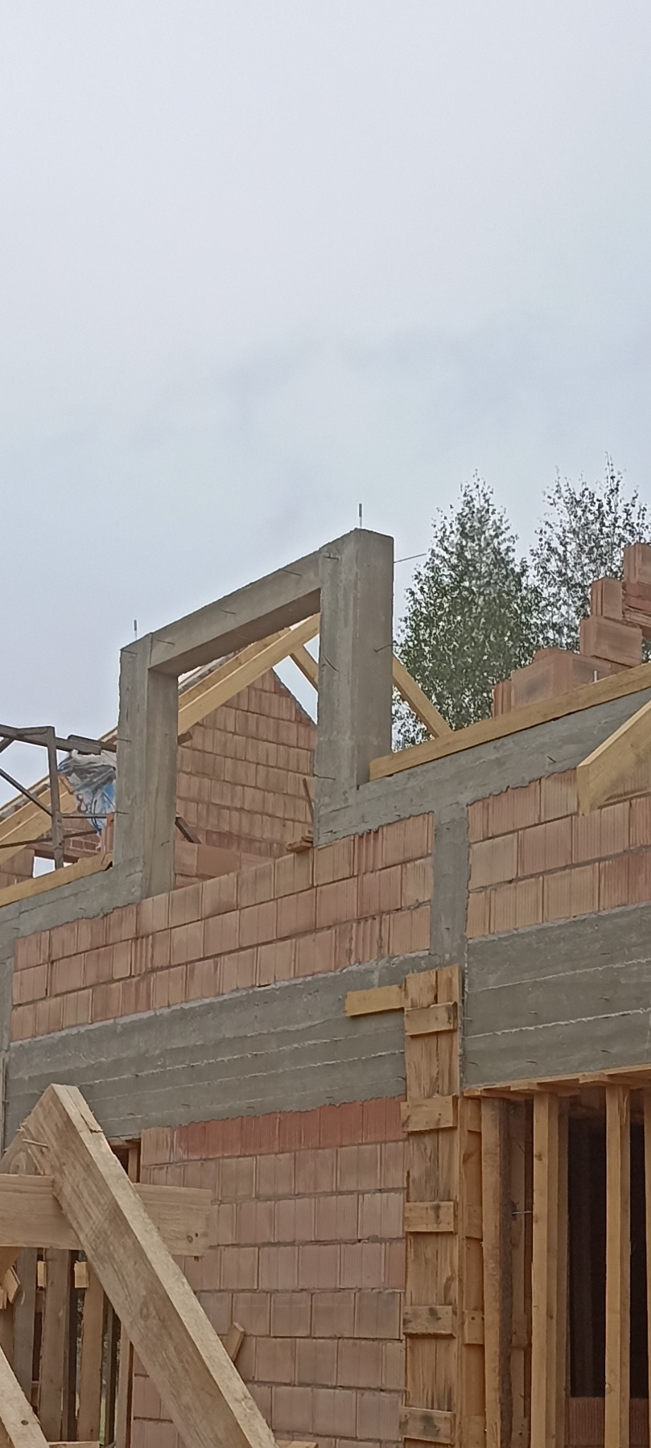 Budowa domu z czerwonej cegły: wzniesione ściany z otworem okiennym, widoczna więźba dachowa i szalunki. Etap surowy otwarty, pochmurne niebo w tle.