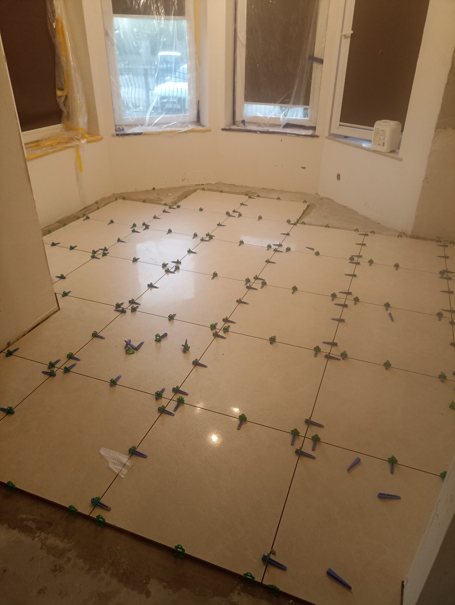 Układanie beżowych płytek podłogowych w pomieszczeniu z oknami, z użyciem zielonych i niebieskich krzyżyków dystansowych. Częściowo ułożona podłoga, widoczne przygotowania.