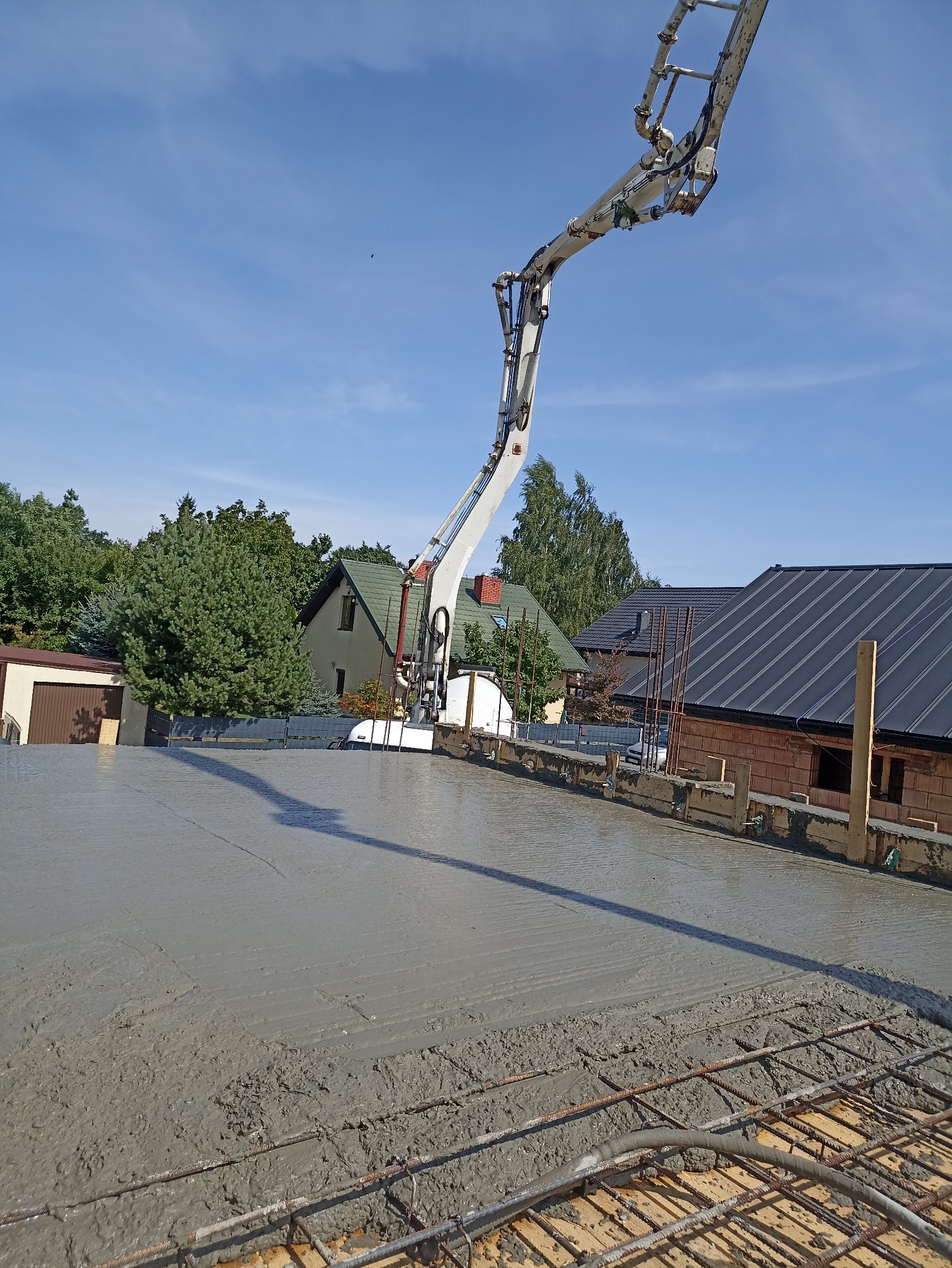 Wylewanie betonu na strop zbrojony, widoczna pompa do betonu. W tle domy jednorodzinne i błękitne niebo. Prace budowlane w toku, etap zalewania stropu.