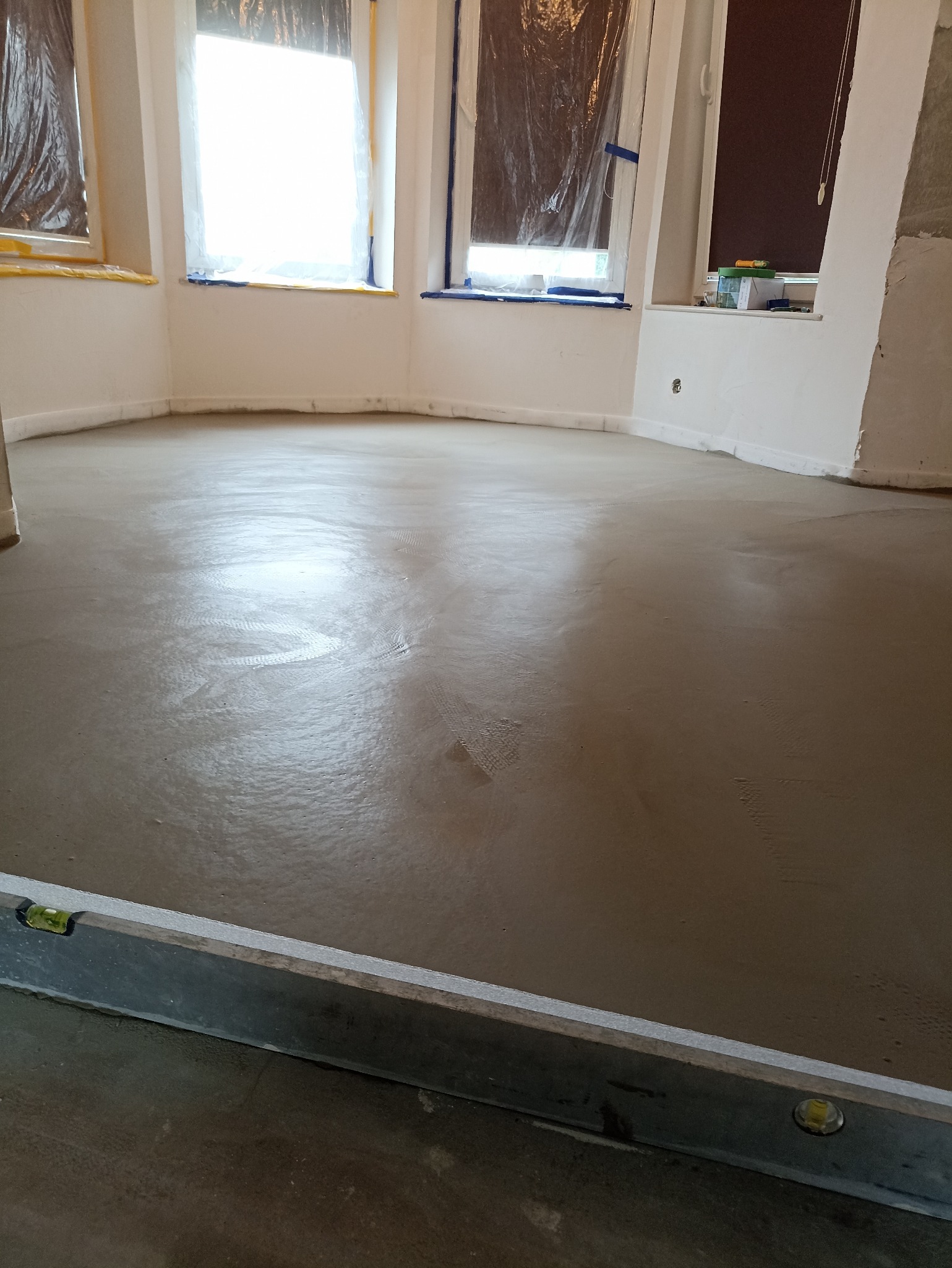 Wylana, świeża wylewka samopoziomująca w jasnym pomieszczeniu z oknami zabezpieczonymi folią. Poziomica w dolnej części kadru. Widoczne ślady po rozprowadzaniu masy.