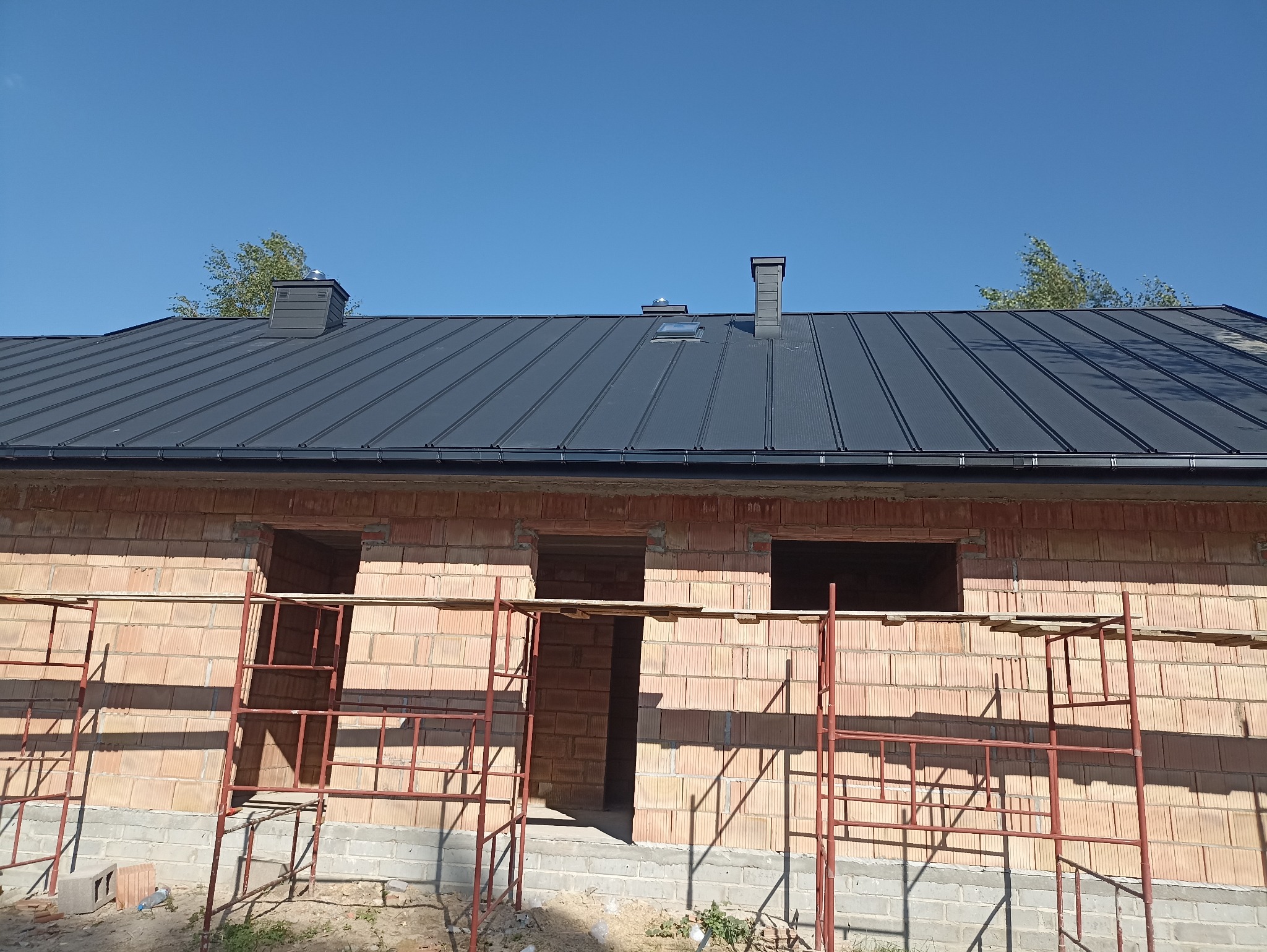Budowa domu z czerwonej cegły z zamontowanym, ciemnym dachem z blachy trapezowej. Widoczne rusztowania i okna bez szyb. Błękitne niebo w tle.