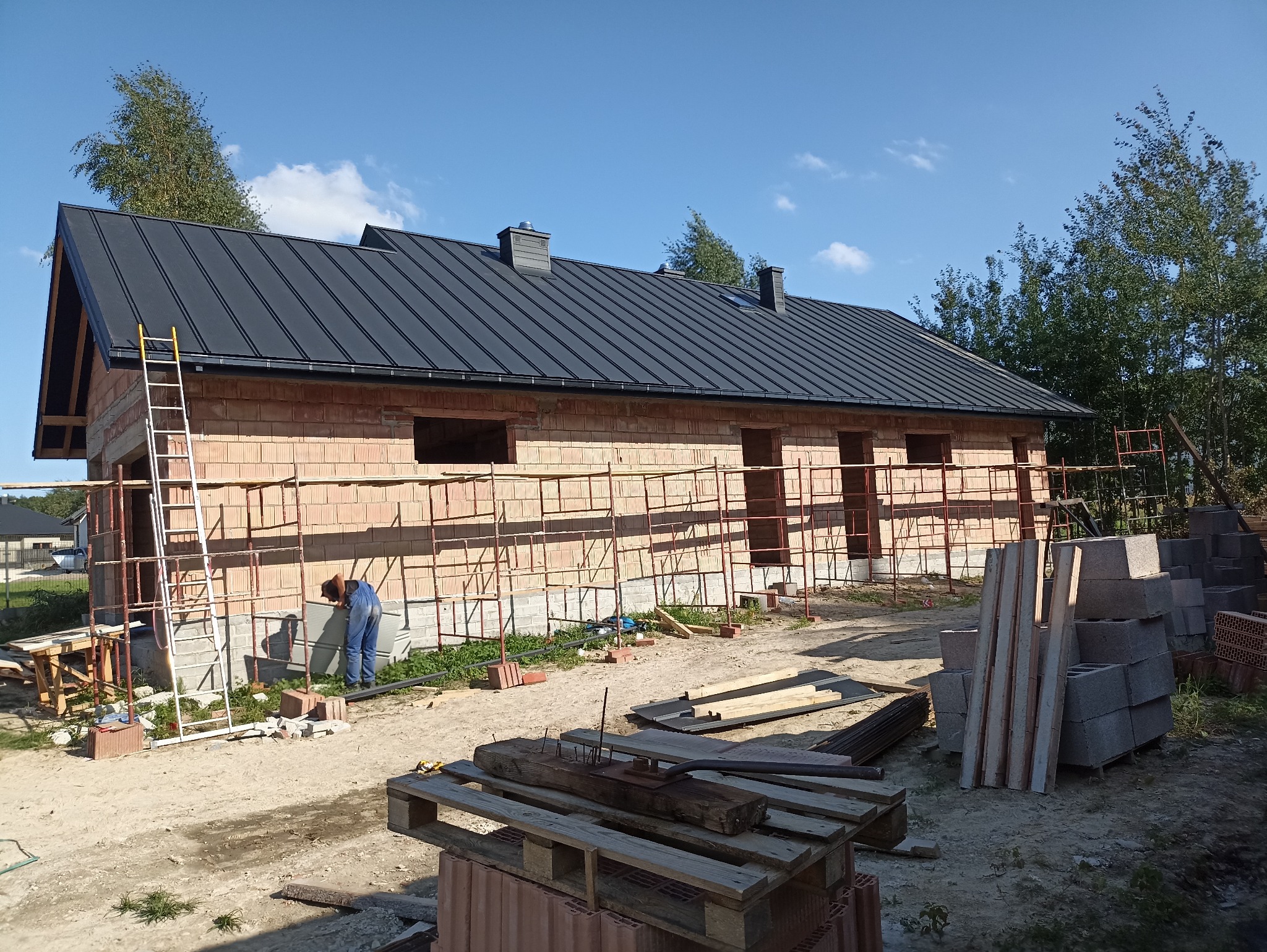 Budowa domu z czerwonej cegły, czarny dach, rusztowania, pracownik w niebieskim kombinezonie przy fundamentach. Widok na plac budowy w słoneczny dzień.