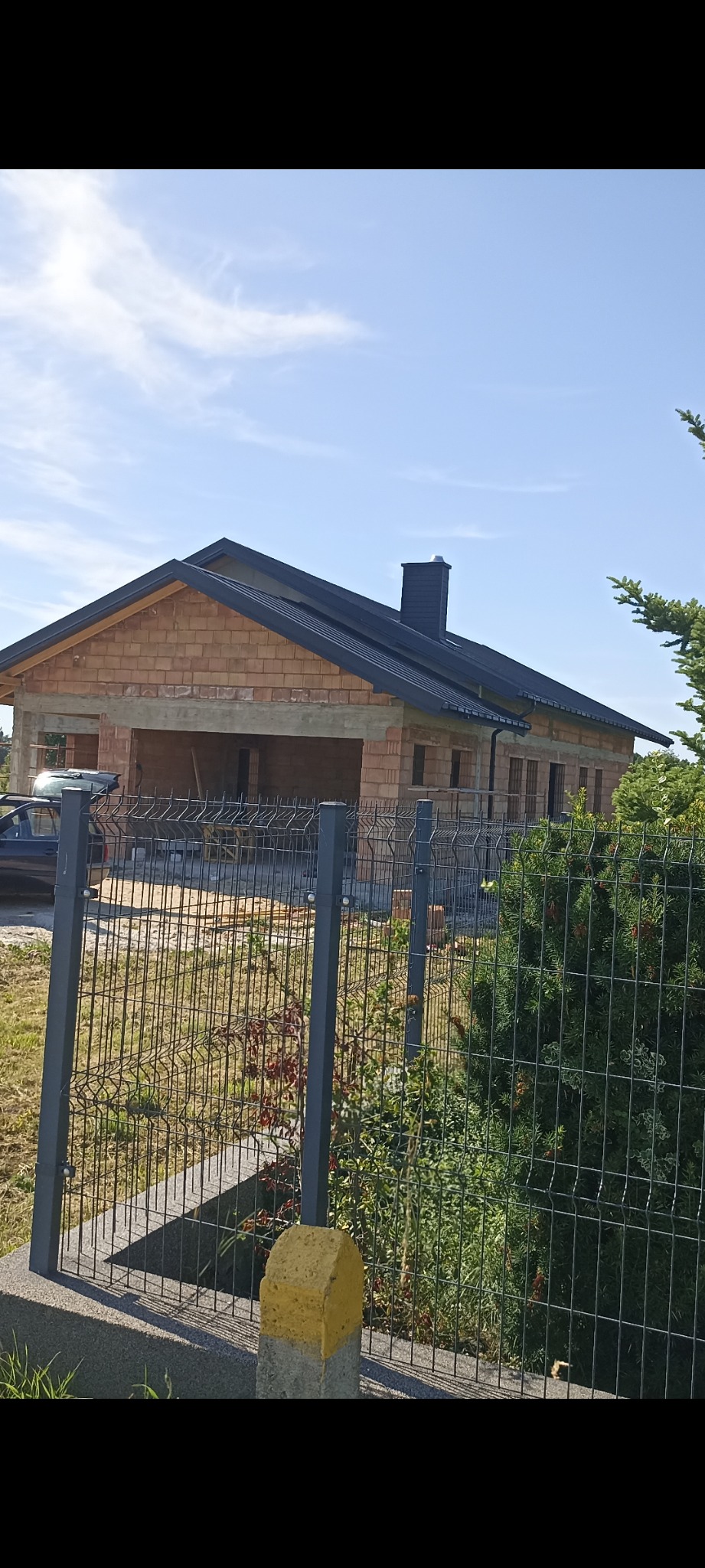 Budowa domu z cegły ceramicznej, widoczna więźba dachowa z czarną blachą. Dom w stanie surowym otwartym, ogrodzony siatką. Słoneczny dzień.
