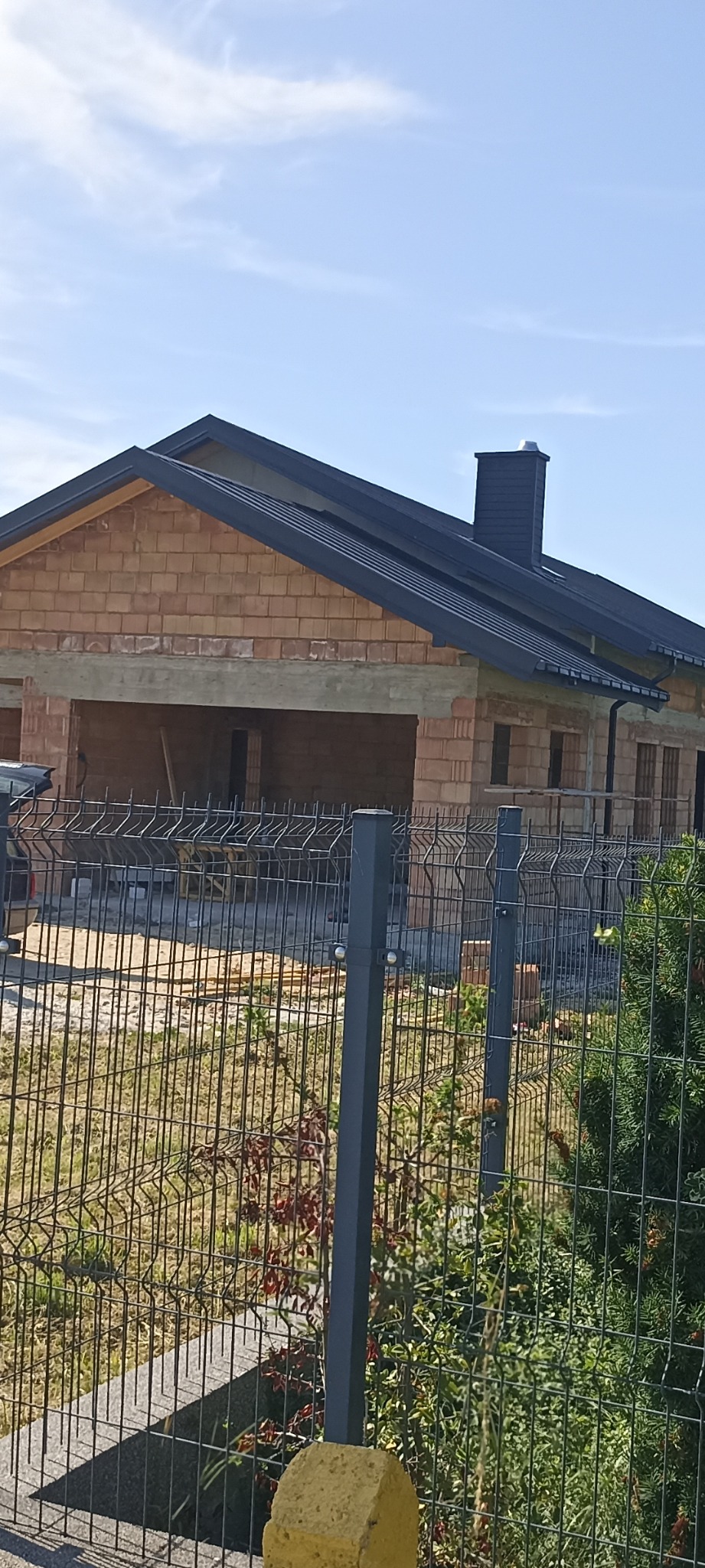 Surowy stan domu murowanego z ciemnym dachem, widoczny przez metalowe ogrodzenie. Brak okien, widoczny komin i garaż. Niebo błękitne, słoneczny dzień.