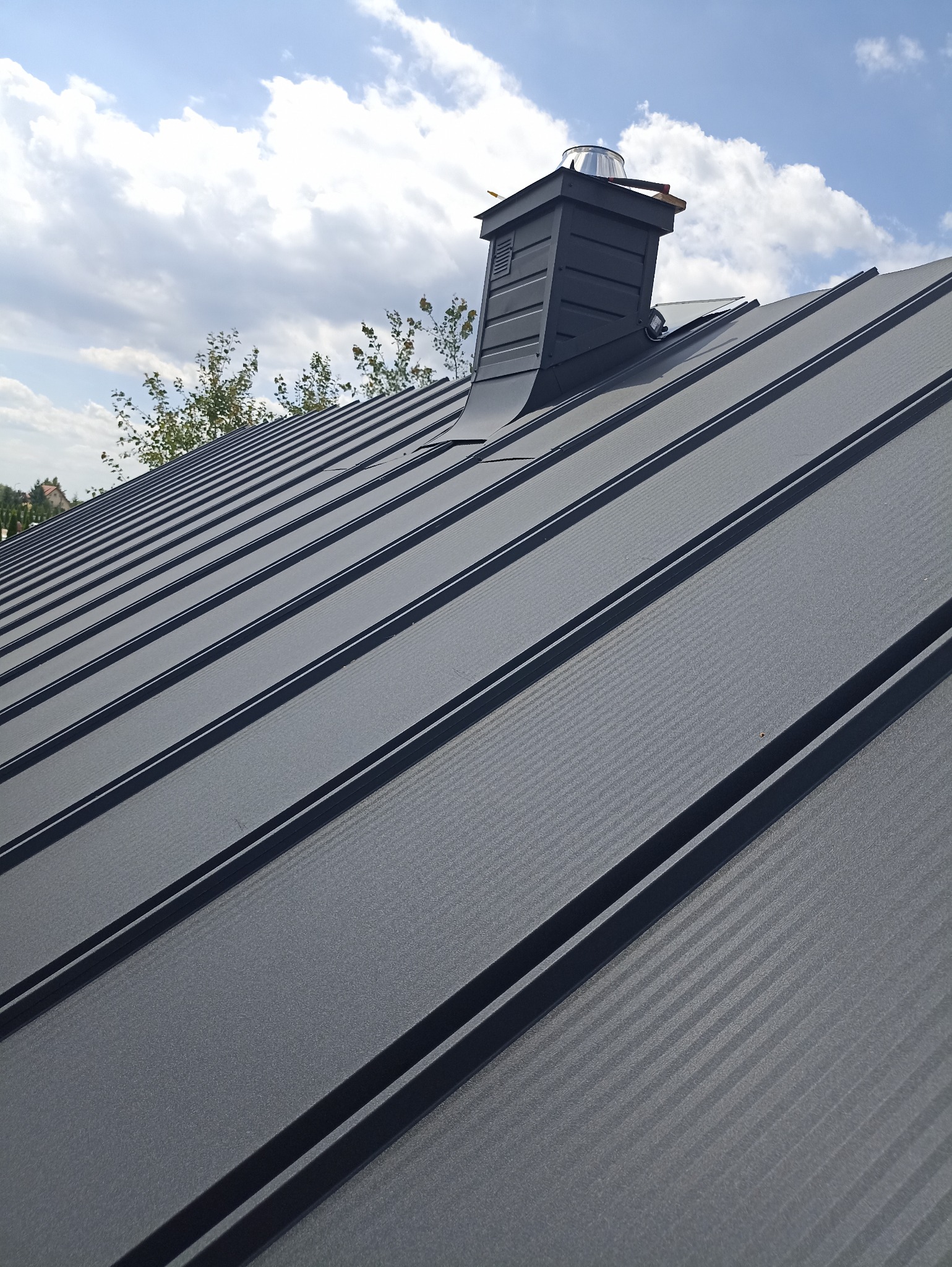 Szary dach z blachy trapezowej z kominem na tle błękitnego nieba z chmurami. Widok z dołu, ukazujący strukturę blachy i detale komina.