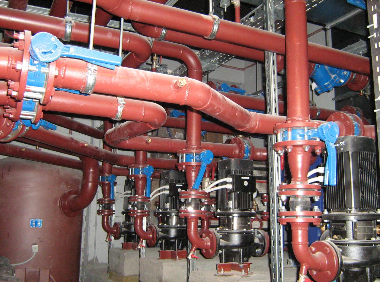 Złożona instalacja z czerwonych rur i niebieskich zaworów w kotłowni. Widoczne pompy obiegowe i zbiornik wyrównawczy. Elementy instalacji przymocowane do ściany.