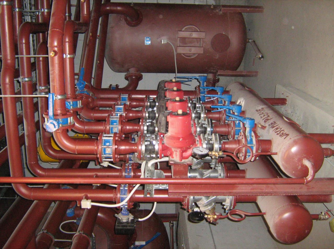 Złożona instalacja rur i zbiorników w odcieniach czerwieni i brązu, z niebieskimi zaworami, widoczny manometr i inskrypcja na zbiorniku.