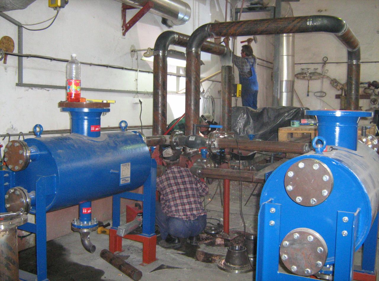 Instalacja przemysłowa w trakcie montażu: spawanie rur, dwa niebieskie zbiorniki ciśnieniowe, butelka z napojem na jednym z nich. Prace instalacyjne w toku.