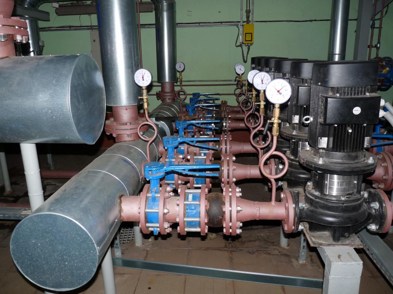 Złożona instalacja przemysłowa z rurami, zaworami z niebieskimi uchwytami i manometrami, zasilana przez czarne silniki, w pomieszczeniu o zielonych ścianach.