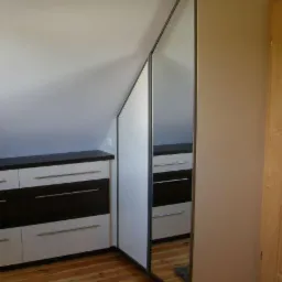 Szafa wnękowa z lustrzanymi drzwiami przesuwnymi, dopasowana do skosu poddasza, obok komoda z szufladami w kolorze białym i brązowym, drewniana podłoga.