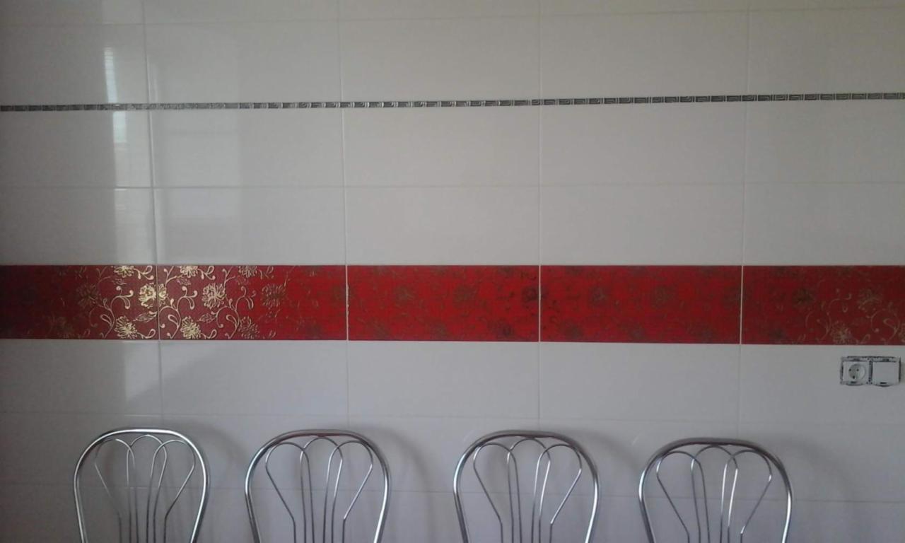 Wykończona ściana z białych płytek, ozdobiona poziomym pasem czerwonych płytek z motywem kwiatowym i cienkim paskiem szarych płytek. Widoczne cztery krzesła.