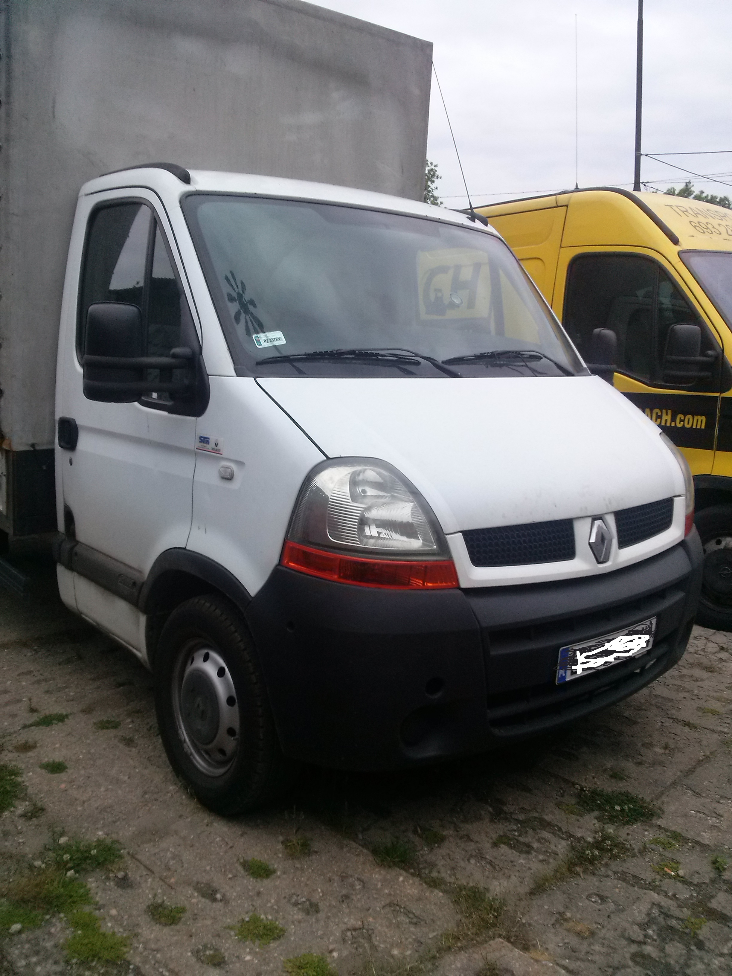 Biały bus Renault Master z plandeką, zaparkowany obok żółtego busa. Widoczna szyba z naklejką i tablica rejestracyjna. Zdjęcie z zewnątrz, w dzień.