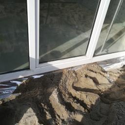 DANIEL MATYSIAK matysbudgw - Przygotowanie podłoża pod taras: piasek podsypany pod oknem tarasowym, folia izolacyjna widoczna pod piaskiem, fragment sylwetki osoby w czapce.