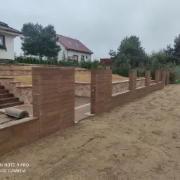 Wznoszenie ogrodzenia z betonowych bloczków w ogrodzie, widoczne także schody z kamienia i przygotowany teren pod trawnik.