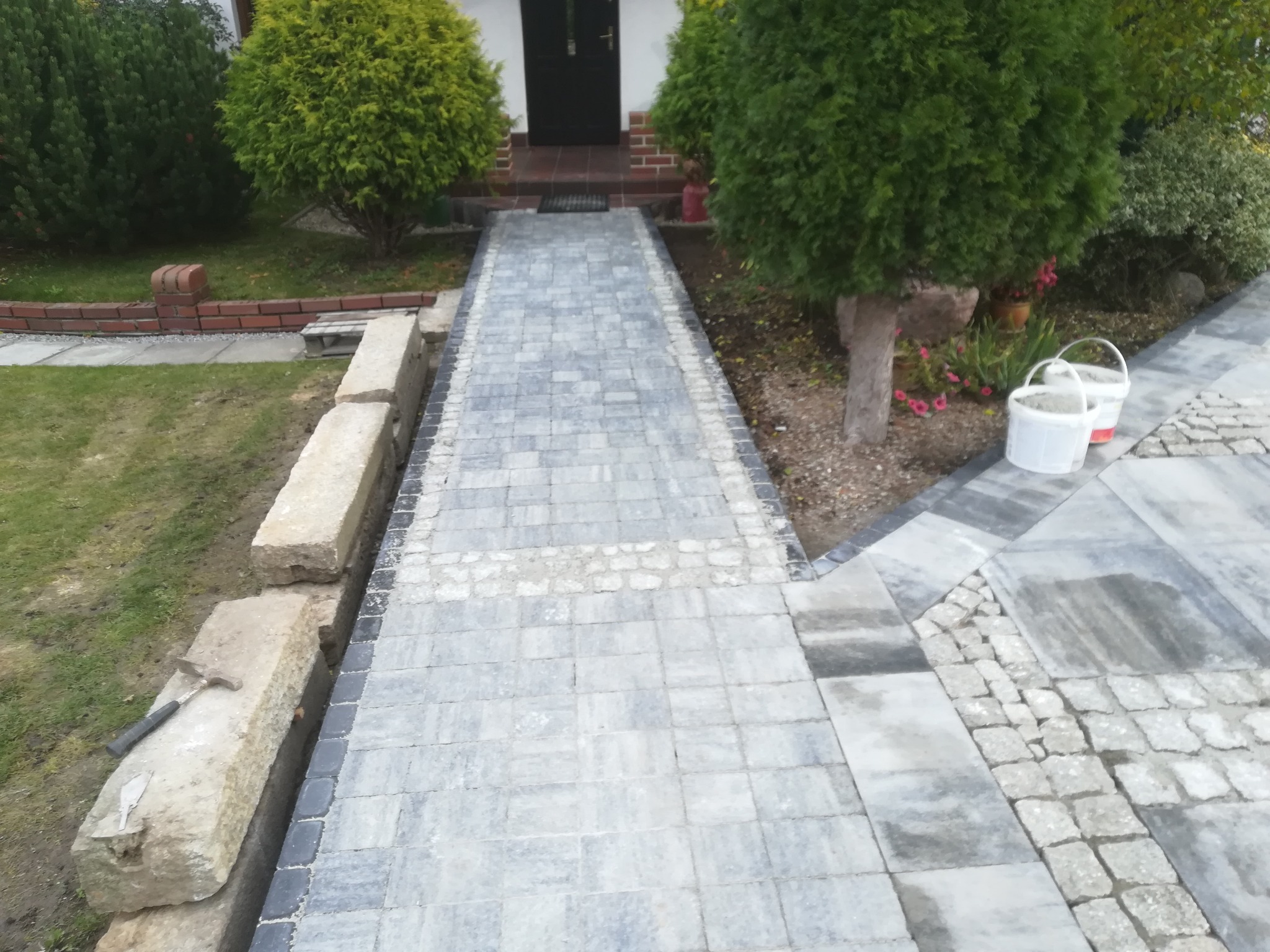Ścieżka z szarej kostki brukowej prowadząca do drzwi wejściowych, obrzeża z jasnego kamienia, widoczne narzędzia brukarskie i białe wiaderka, otoczenie zielone krzewy.