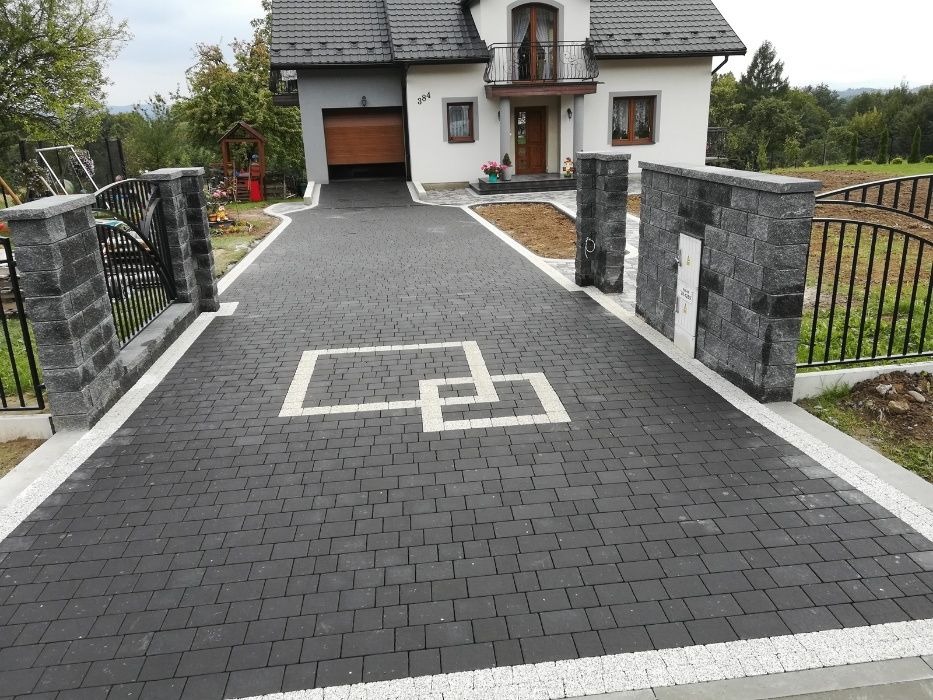 Czarna kostka brukowa z białym wzorem geometrycznym na podjeździe, obrzeża z jasnego kamienia, brama z elementami z szarej kostki i metalu, dom z garażem w tle.