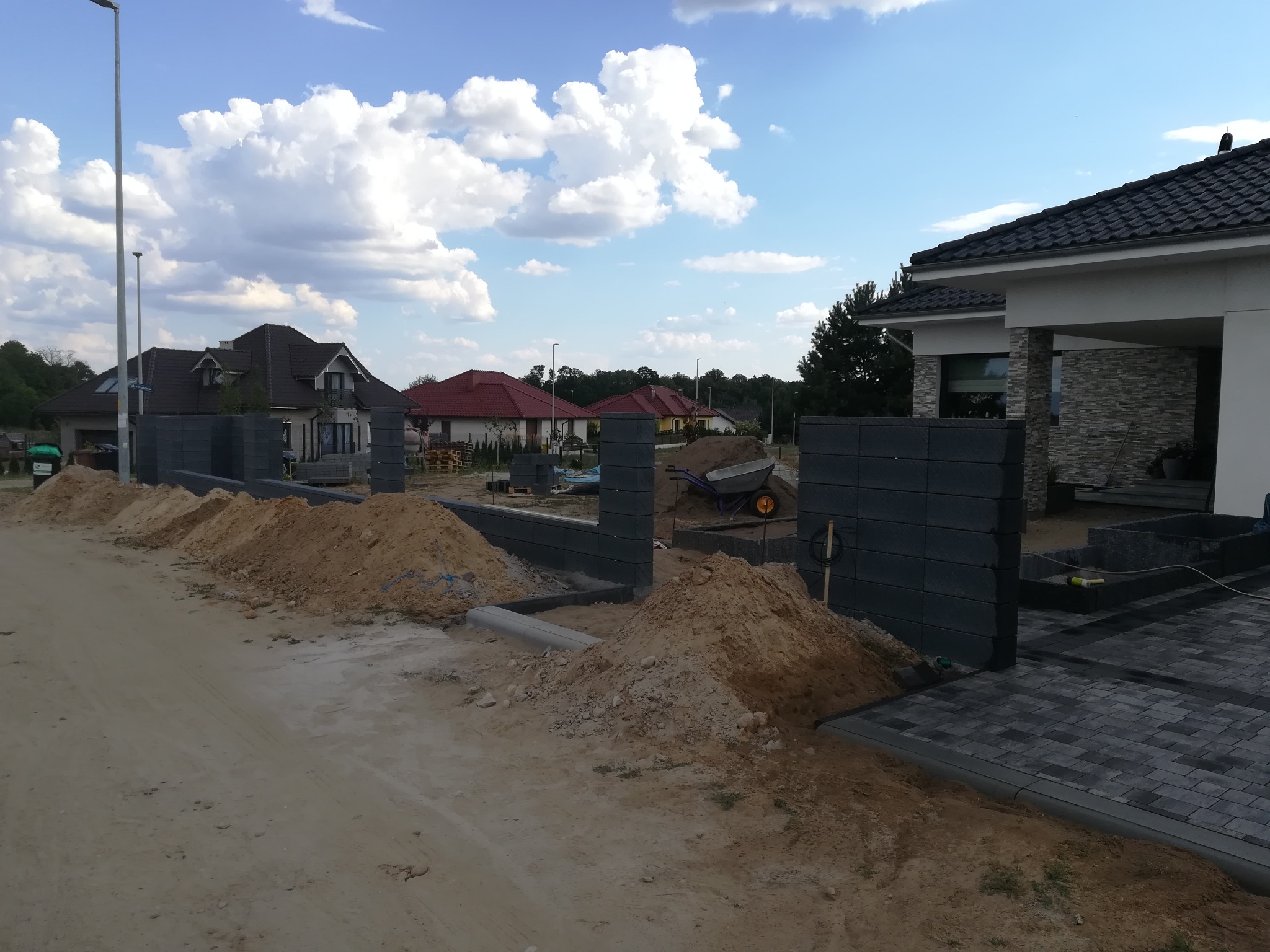 Budowa ogrodzenia z betonowych bloczków i układanie kostki brukowej na podjeździe, widoczne hałdy piasku i narzędzia pracy.