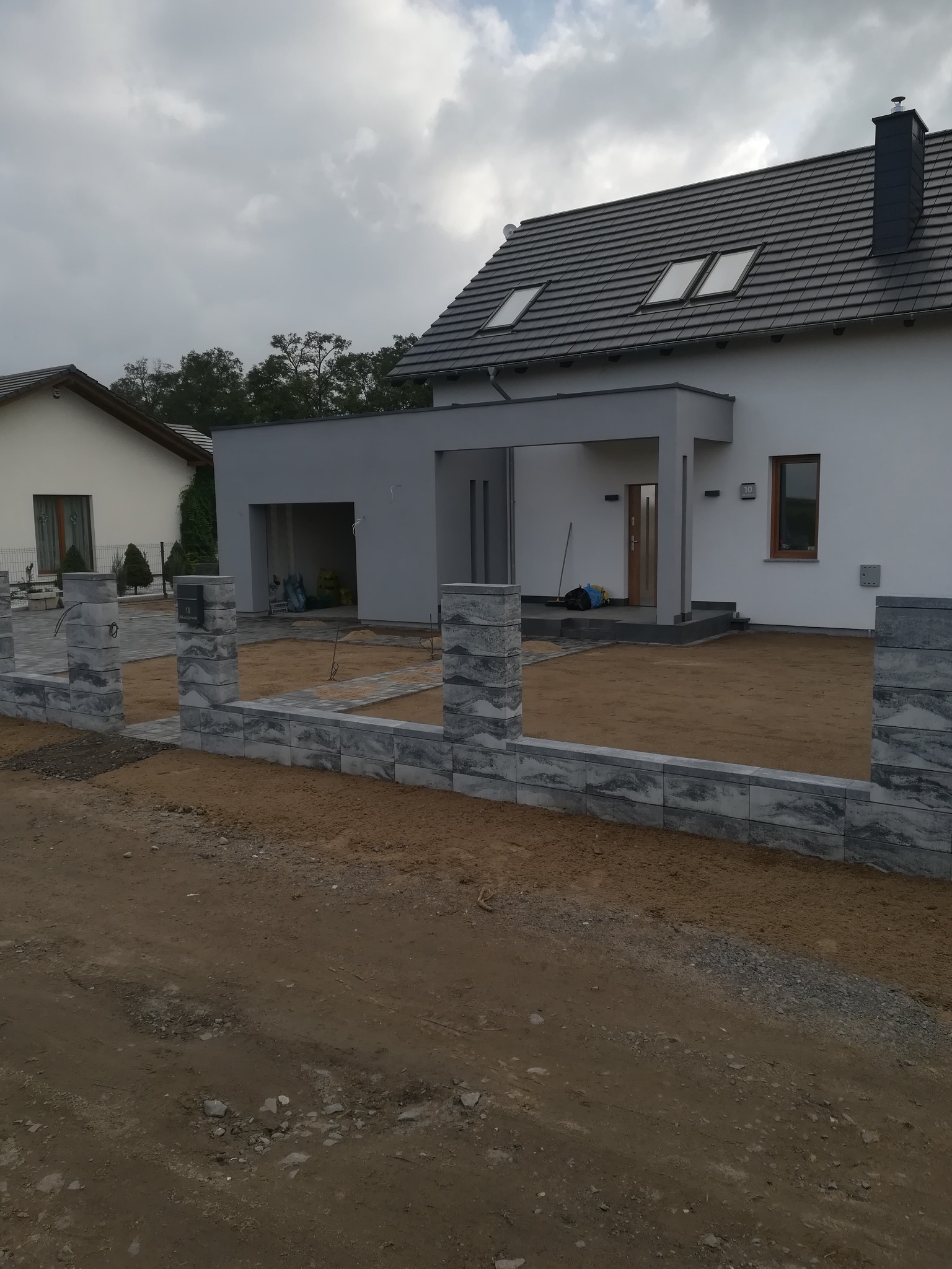 Wznoszone ogrodzenie z szarej kostki betonowej o marmurkowym wzorze, widoczny fragment posesji z przygotowanym podłożem i częściowo ułożoną ścieżką z kostki, dom w tle pod pochmurnym niebem.