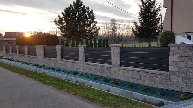 Nowoczesne ogrodzenie z poziomych paneli w kolorze grafitowym osadzone na murze z szarej kostki łupanej, widoczne nasadzenia wzdłuż ogrodzenia, w tle dom i zachodzące słońce.
