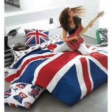 Młoda kobieta gra na czerwonej gitarze elektrycznej siedząc na pościeli z flagą Wielkiej Brytanii. W tle widoczne poduszki i płyty winylowe.