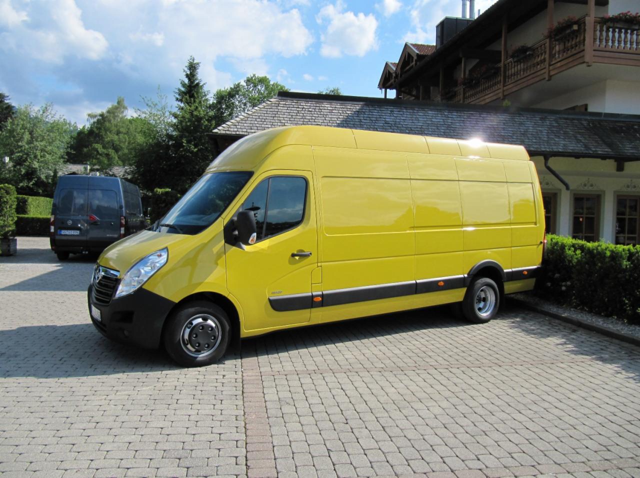 Żółty van Opel Movano na brukowanym podjeździe, z szarym vanem w tle, przed budynkiem w stylu bawarskim. Słoneczny dzień, błękitne niebo.