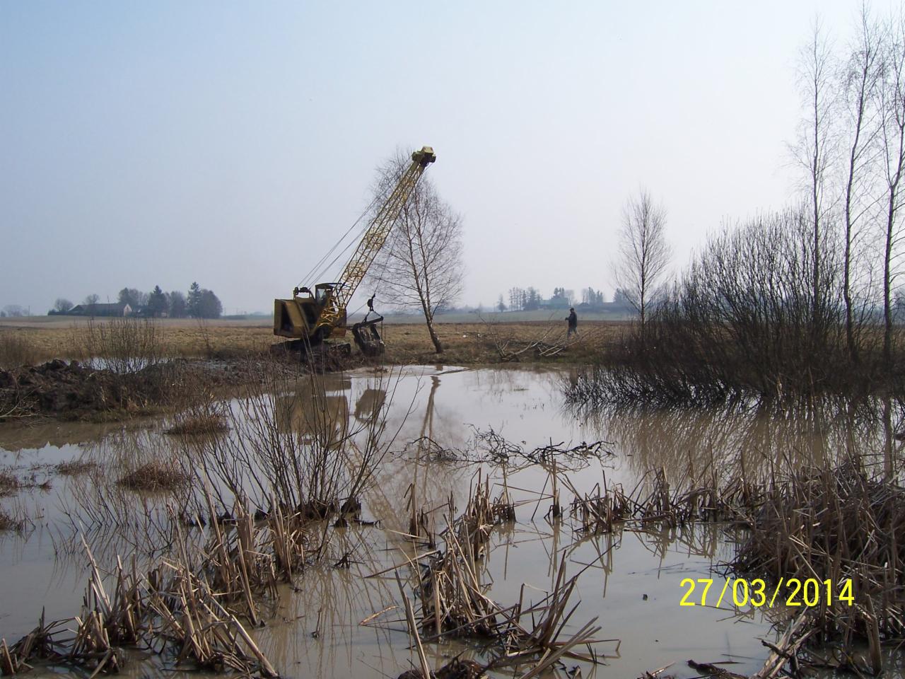 Żółty dźwig w podmokłym terenie, odbicie w wodzie, w tle budynki i samotna postać. Data 27/03/2014 w prawym dolnym rogu.