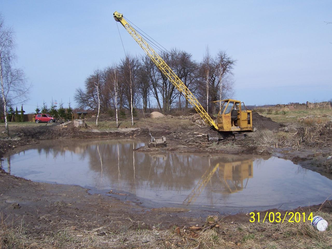 Żółty dźwig nad wykopem wypełnionym wodą, odbicie w lustrze wody. W tle drzewa i czerwony samochód. Data 31/03/2014 w prawym dolnym rogu.