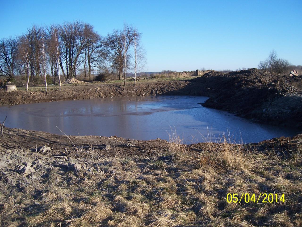 Wykonanie stawu z folią w Suwałkach. Widok na wykopany zbiornik wodny z folią na dnie, otoczony ziemią i trawą, pod błękitnym niebem. Data wykonania: 05/04/2014.