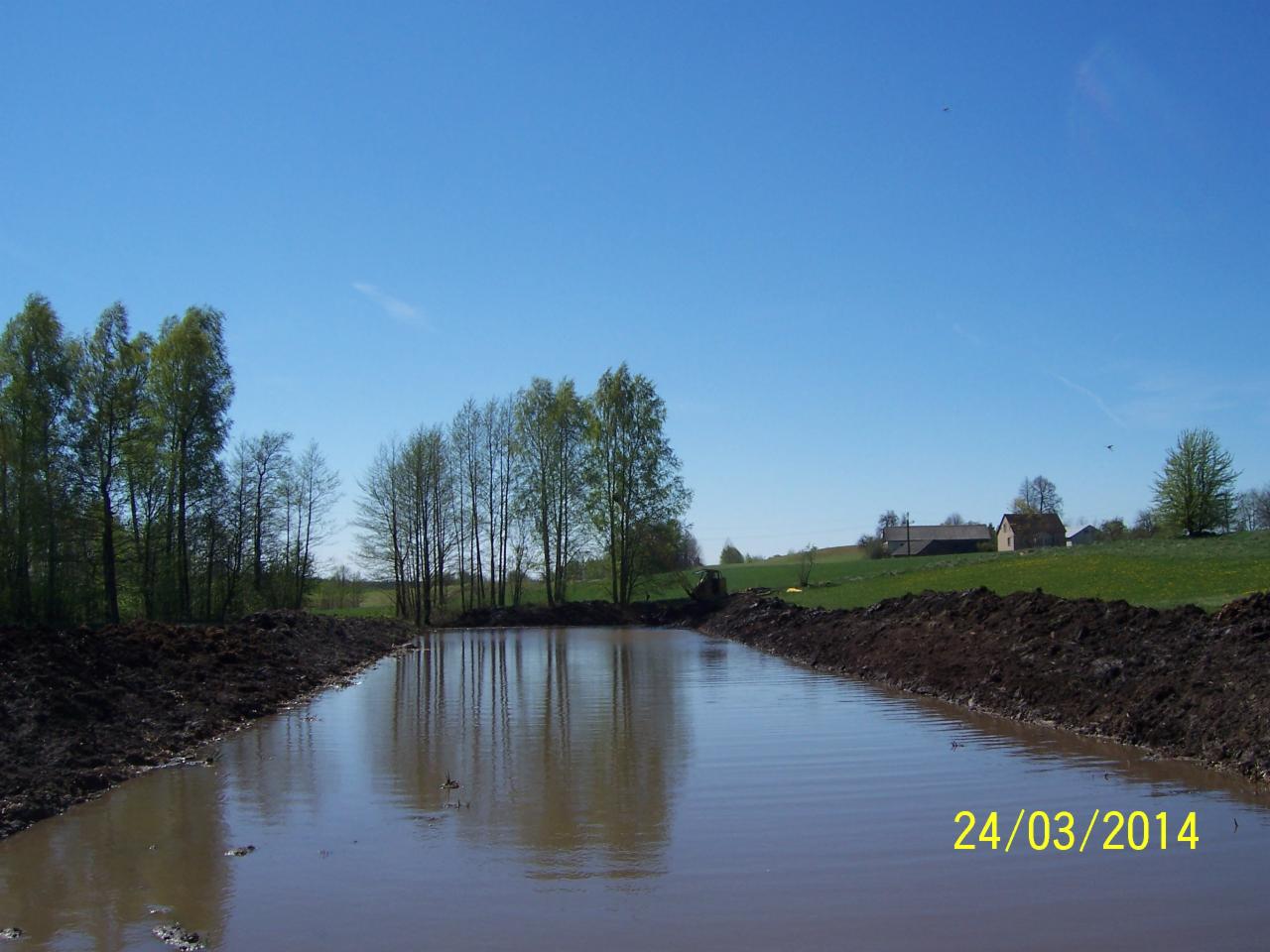 Wykop pod zbiornik wodny z brzegami umocnionymi ziemią. W tle drzewa i budynki. Data 24/03/2014 w prawym dolnym rogu.