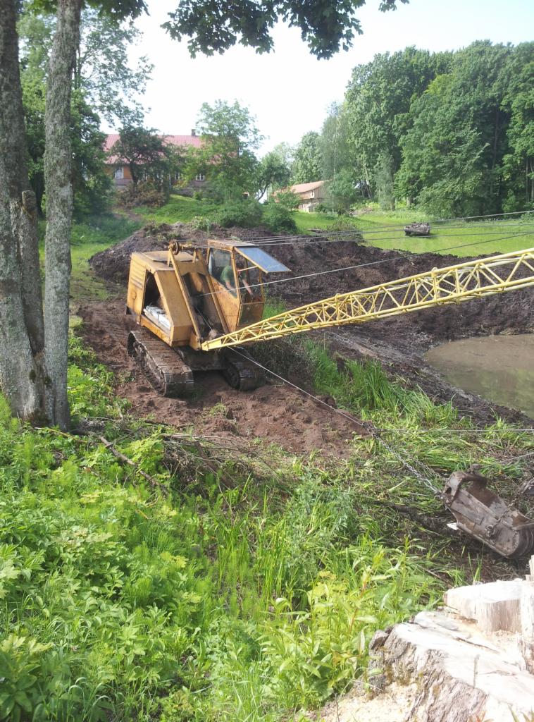 Żółty dźwig gąsienicowy na nierównym terenie, prawdopodobnie w trakcie prac ziemnych przy zbiorniku wodnym. W tle widoczny budynek i drzewa.