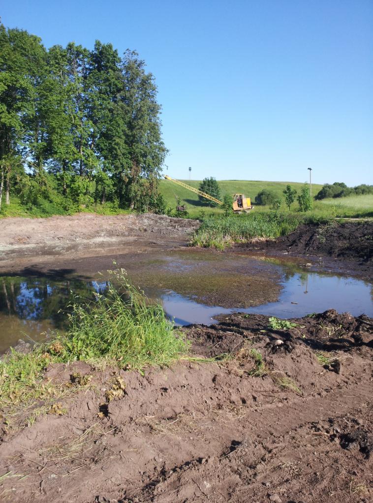 Żółta koparka na tle zielonego wzgórza, częściowo zanurzona w wodzie. Na pierwszym planie rozkopana ziemia, ślady opon i roślinność.