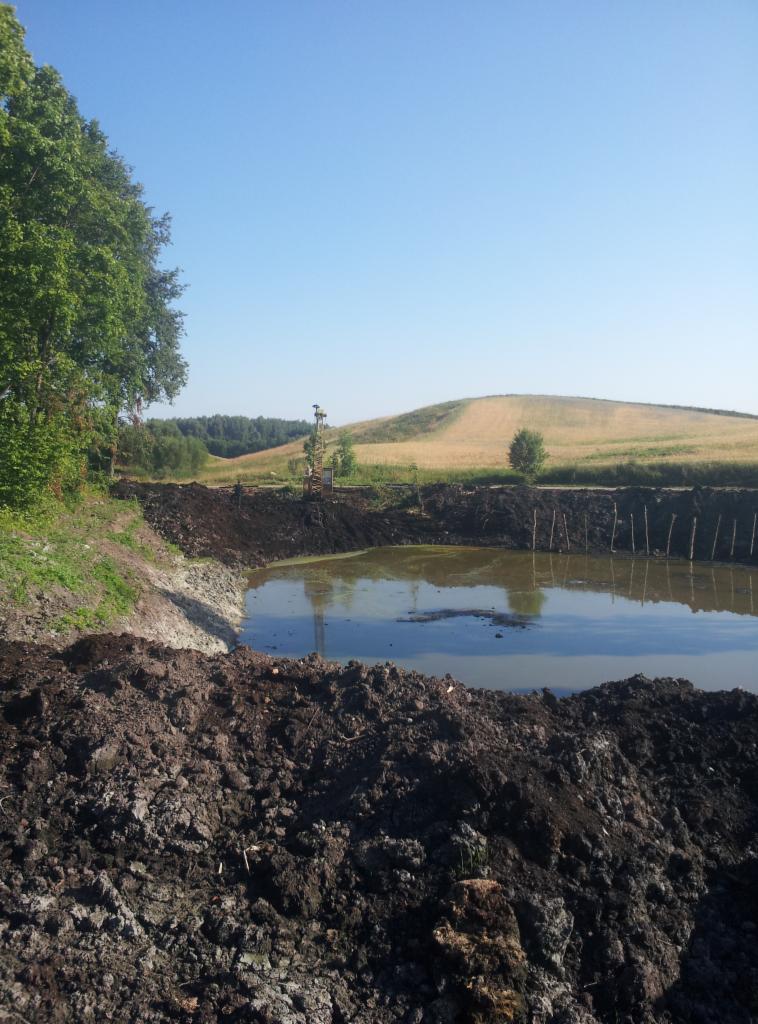 Wykopany staw w ziemi z palikami brzegowymi i zielonym wzgórzem w tle, wykonany w słoneczny dzień. Prace ziemne.