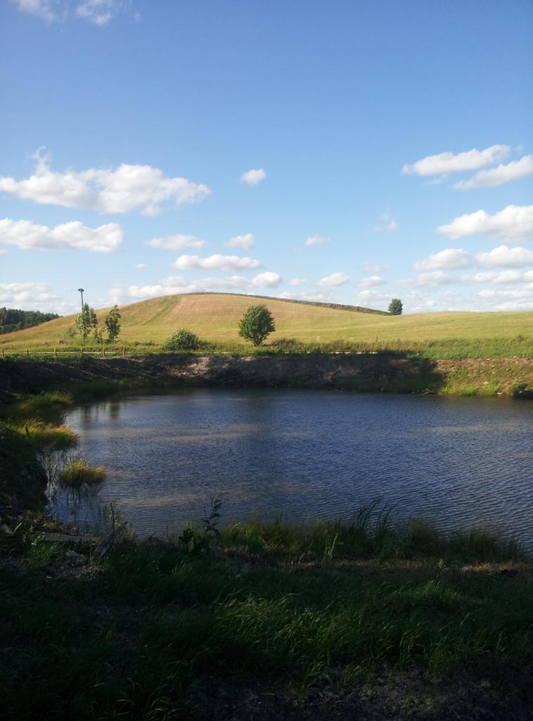 Malowniczy krajobraz z oczkiem wodnym na pierwszym planie, wzgórzem w tle i błękitnym niebem z chmurami. Woda lekko pofalowana. Ujęcie w słoneczny dzień.