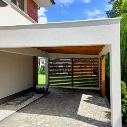 Nowoczesna wiata garażowa z białymi ścianami i drewnianą konstrukcją, widoczne szare kostki brukowe i zieleń w tle.