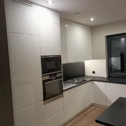 Nowoczesna kuchnia z białymi, lakierowanymi frontami, czarnym blatem i wbudowanym piekarnikiem oraz płytą gazową, oświetlona punktowymi lampami sufitowymi, z widoczną drewnianą podłogą.