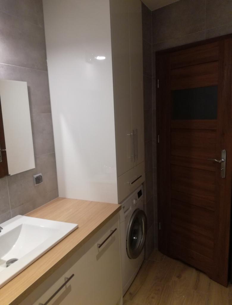 Łazienka z zabudową meblową: biała szafa łazienkowa z połyskiem, blat z jasnego drewna, umywalka, pralka w zabudowie i drewniane drzwi w tle.