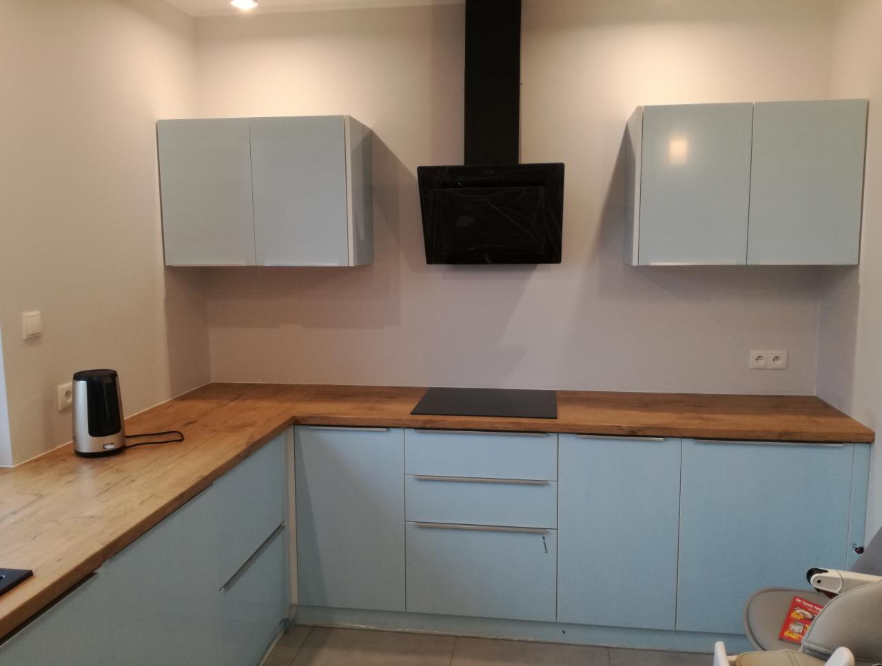 Jasna kuchnia z drewnianym blatem i czarnym okapem, widoczne górne szafki i część zabudowy dolnej, z eksposem na fakturę blatu.