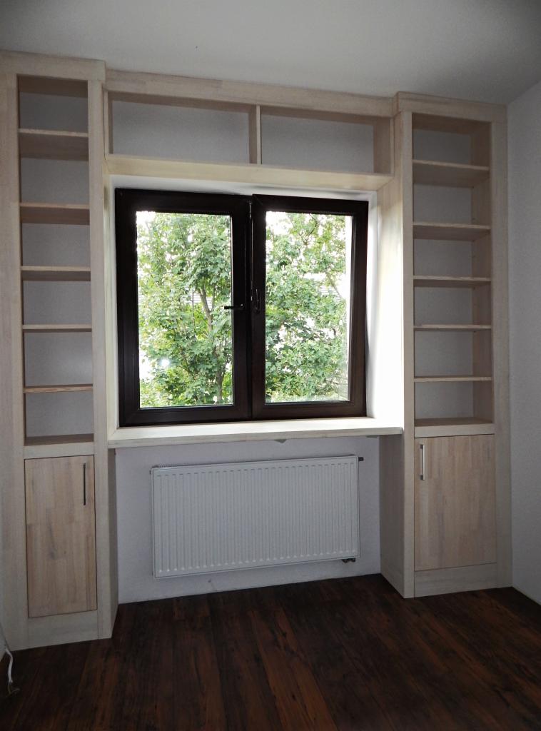 Zabudowa okienna z półkami i szafkami po bokach, wykonana z jasnego drewna, z widokiem na zieleń za oknem.