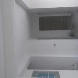 Wnętrze po malowaniu: gładkie, białe ściany i sufit, widoczne otwory na gniazdka, fragment okna z niebieskimi szybami i sprzęt w tle, ukazujący etap wykończeniowy.