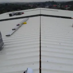 Biały, blaszany dach z widocznymi łączeniami, na którym leżą materiały budowlane w folii, profile aluminiowe i puste kartony, z widokiem na zabudowania w tle.