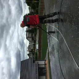 Pracownik w czerwonej kamizelce z logo firmy czyści powierzchnię betonową myjką ciśnieniową na tle pochmurnego nieba i zabudowań mieszkalnych.