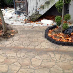 Nowo ułożona, wzorzysta kostka brukowa prowadząca do schodów zewnętrznych, z obrzeżami rabat, w tle widoczne materiały budowlane i narzędzia.