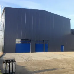 Duży, ciemnoszary budynek przemysłowy z blachy trapezowej, z niebieskimi bramami segmentowymi i drzwiami wejściowymi, zewnętrzna drabina ewakuacyjna, plac wyłożony kostką betonową, zbiornik...