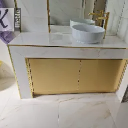 Nowoczesna szafka łazienkowa z białym blatem imitującym marmur, złotymi akcentami i umywalką nablatową; złota armatura, lustro w tle, płytki imitujące marmur na ścianach i podłodze.