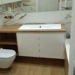 Nowoczesna łazienka z drewnianym blatem pod umywalką, białą szafką, podwieszaną toaletą i marmurową ścianą w tle.