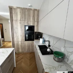 Nowoczesna kuchnia z drewnianymi frontami szafek i białym blatem imitującym marmur, czarny zlew i bateria, parkiet ułożony w jodełkę, widoczne naczynia w zlewie i logo firmy meblowej w rogu.