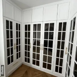 Biała szafa wnękowa z przeszklonymi drzwiami i złotymi uchwytami, widoczny parkiet ułożony w jodełkę.