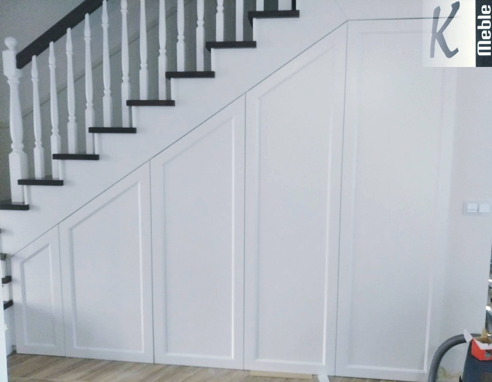 Biała szafa wnękowa pod schodami z drewnianymi stopniami i balustradą, widoczny fragment odkurzacza, logo firmy w prawym górnym rogu.