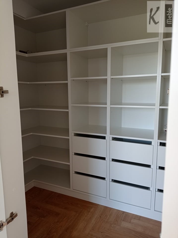 Biała szafa wnękowa z półkami i szufladami, widok z boku, na podłodze parkiet ułożony w jodełkę, w prawym górnym rogu widoczne logo firmy.