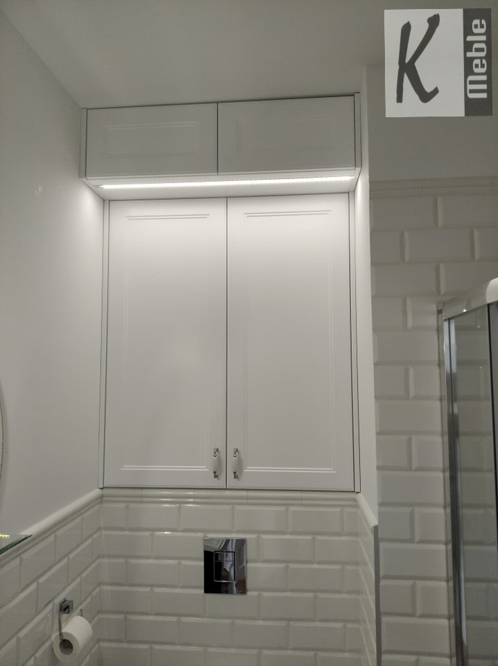 Biała szafka łazienkowa z podświetleniem LED, zamontowana nad toaletą, białe płytki metra w tle, widoczny fragment kabiny prysznicowej.